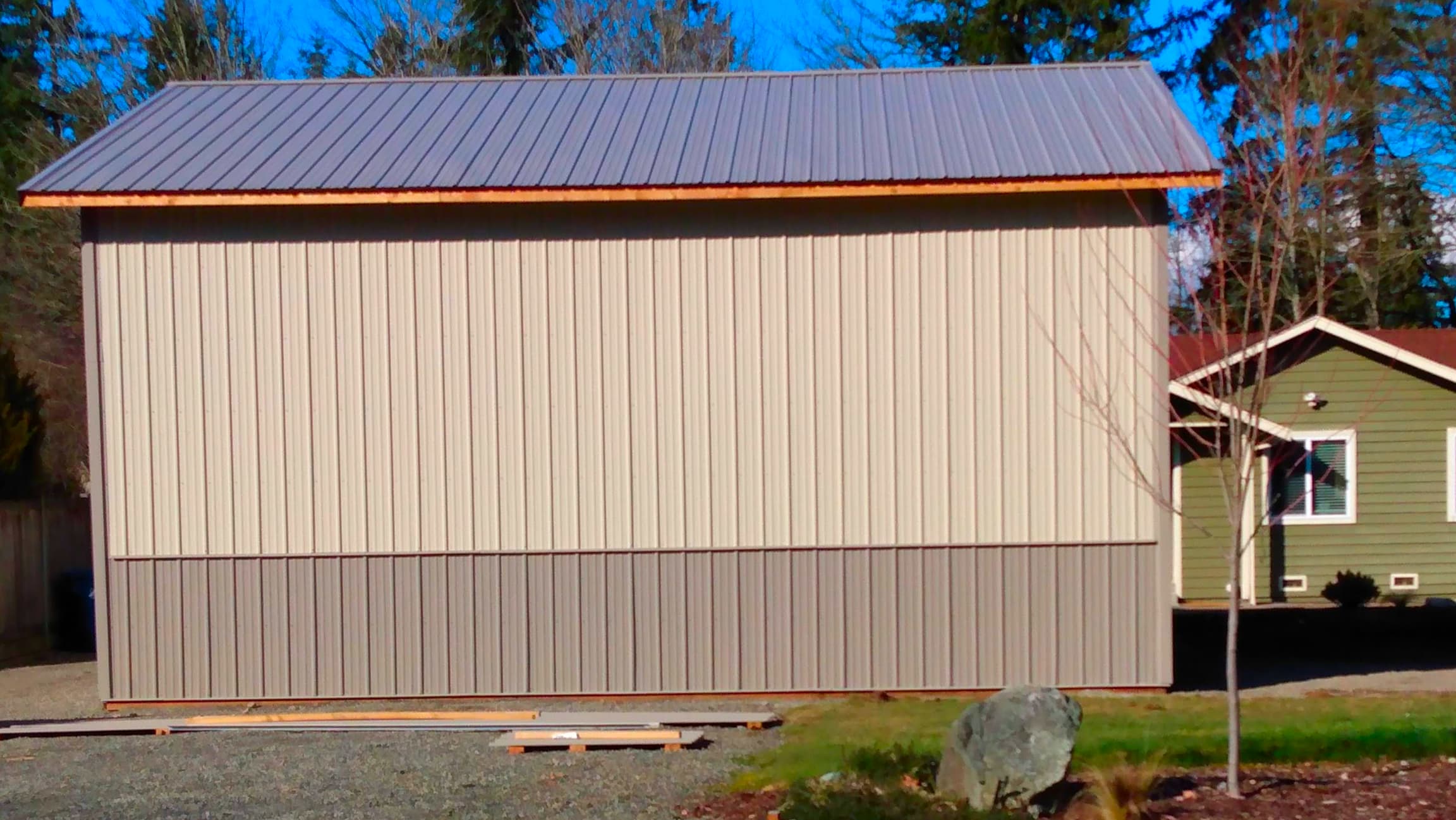 A newly constructed 24'x36' metal storage building by Probuilt Metal Buildings featuring light stone vertical siding with ash grey wainscoting. The building showcases a clean design with metal roof and is set against evergreen trees in Auburn, WA. Perfect for vehicle storage, workshop space, or equipment protection in the Pacific Northwest climate.