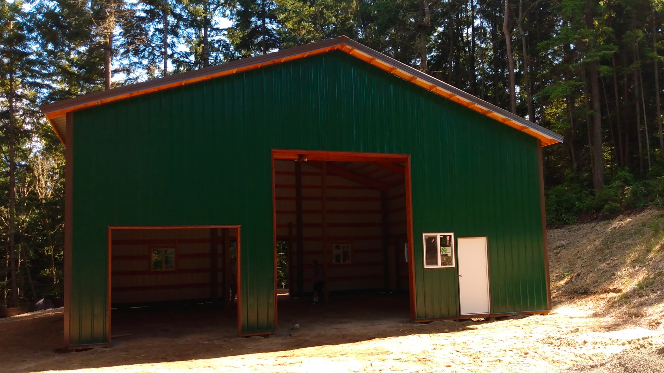 Finished 36'x36' Probuilt Metal building in Olalla featuring forest green metal siding, burnish slate roof, and 18-inch overhangs. This 16' tall structure includes 6 windows, 2 walk-in doors, and 3 overhead doors set against a beautiful Pacific Northwest forest backdrop.